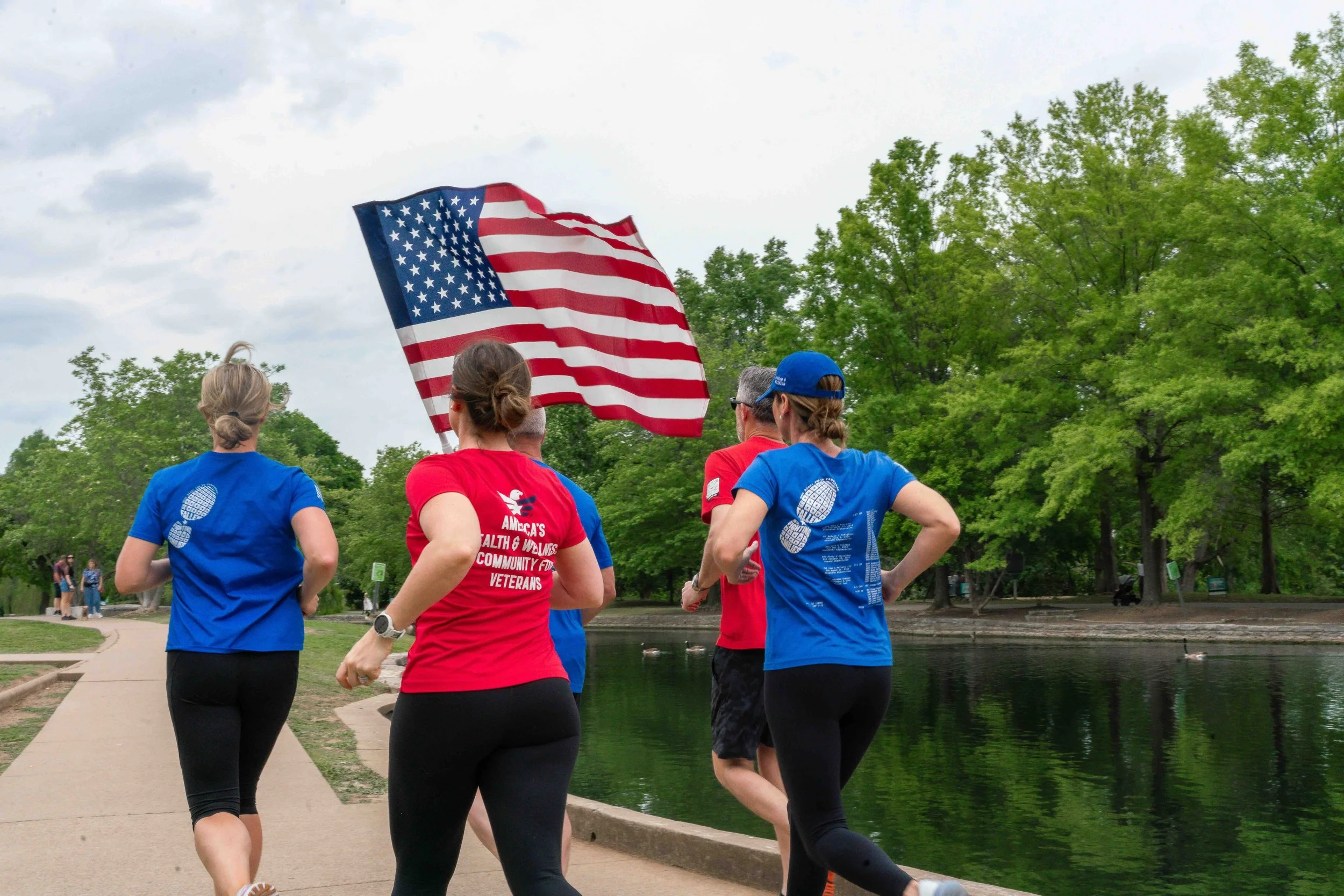 People participating in a run or marathon, some wearing red and blue shirts, running along a park path near a body of water, with an American flag being held up by one participant, surrounded by green trees and a cloudy sky.