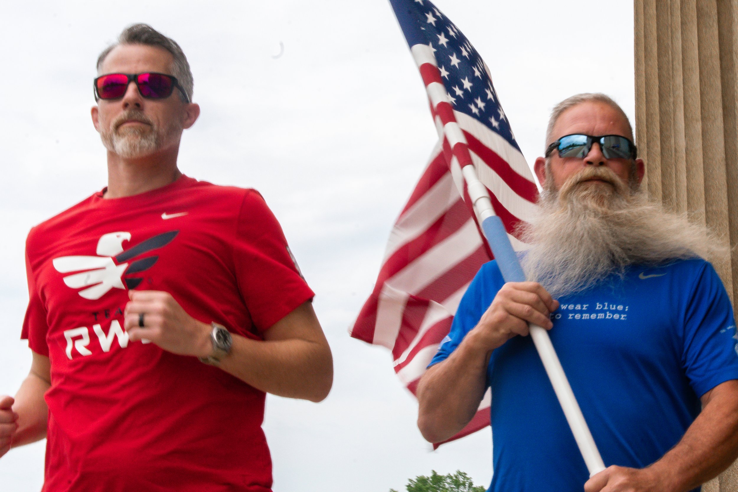 two men running one is wearing a red team rwb shirt and the other a blue wear blue run to remember shirt. the one in the blue shirt is carrying an american flag.