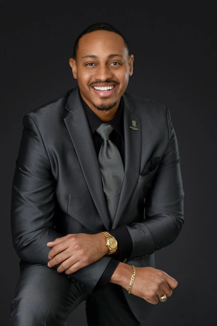A professional man with a dark suit, black shirt, grey tie, and gold watch, smiling against a dark background.