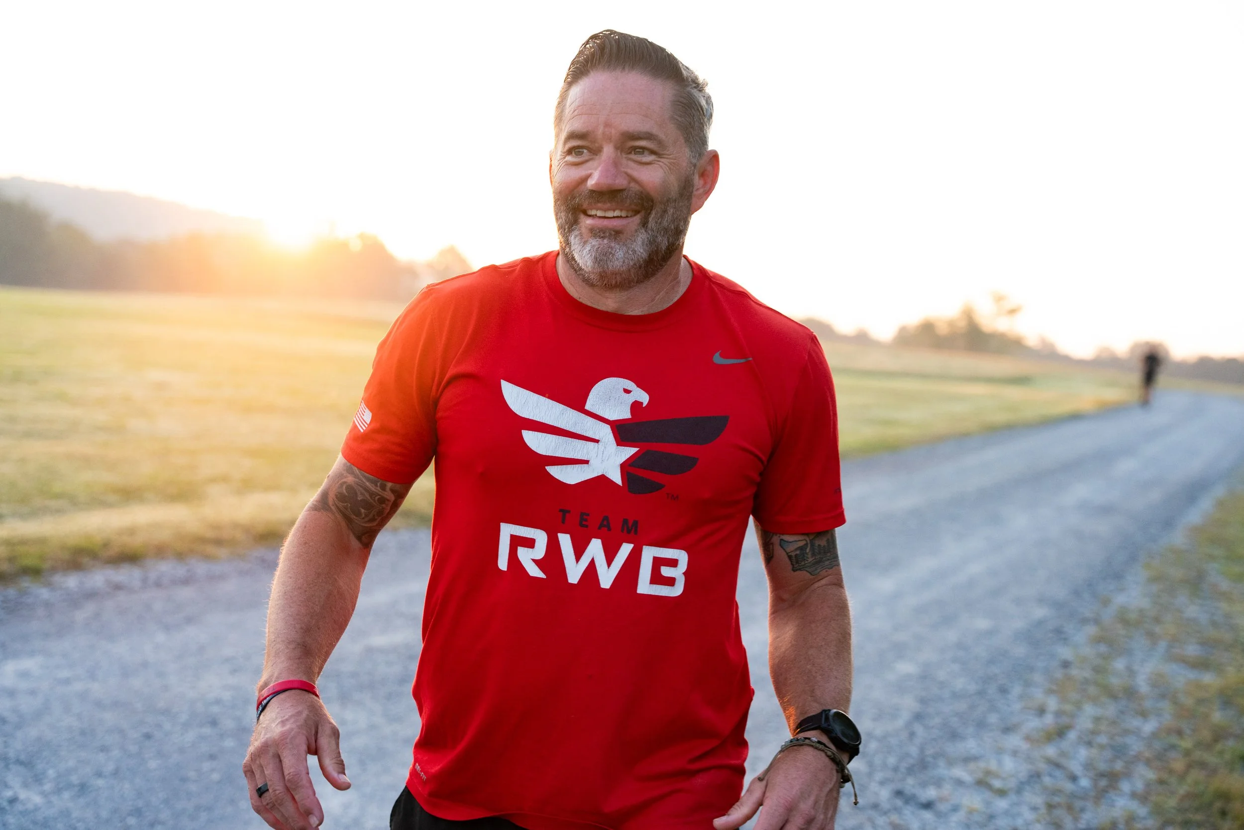 A smiling man with a beard and tattooed arms wearing a red T-shirt with a logo and text, walking outdoors during sunset.
