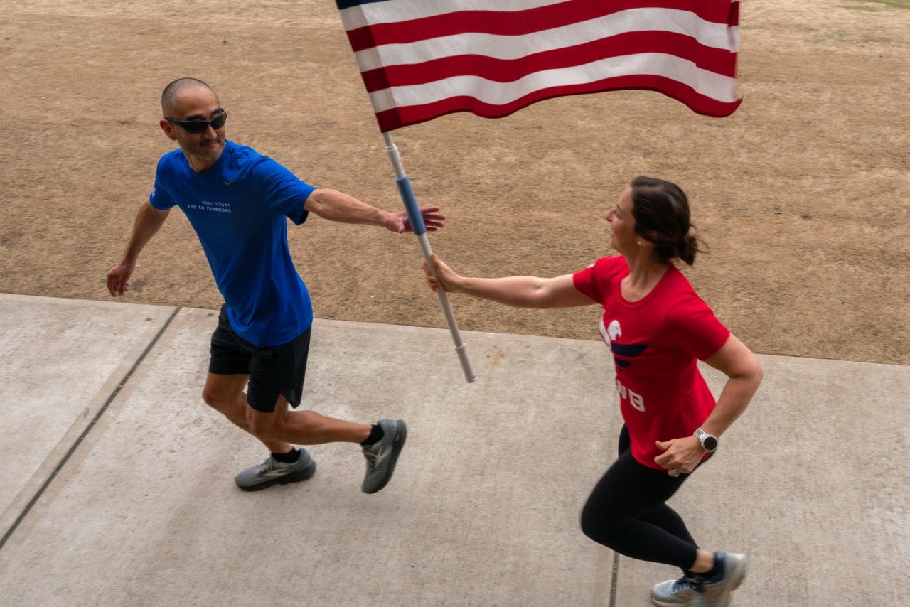 A woman in a red shirt and a man in a blue shirt running outdoors while the woman hands him an American flag.