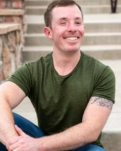 A smiling young man sitting on outdoor steps with a green t-shirt and tattoos on his left arm.