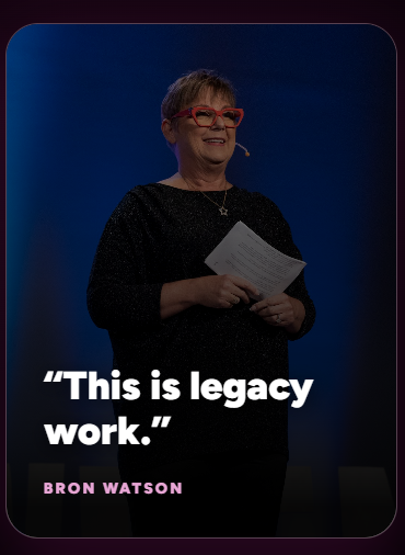 An older woman with short gray hair wearing red glasses and a black sparkly top, holding papers in her hands and smiling on a webinar or conference stage against a blue background.
