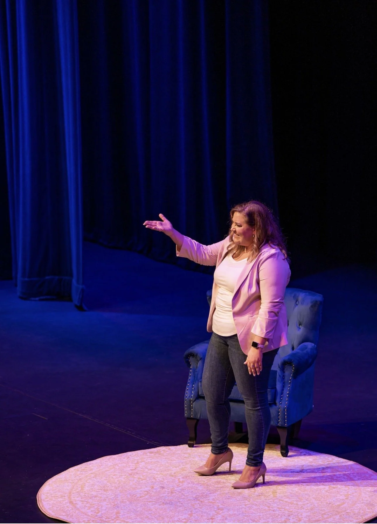 A woman standing on a pink circular rug on stage, smiling, gesturing with her right hand, dressed in a pink blazer, white shirt, and jeans, with a blue velvet armchair behind her, blue stage curtains in the background, under stage lighting.