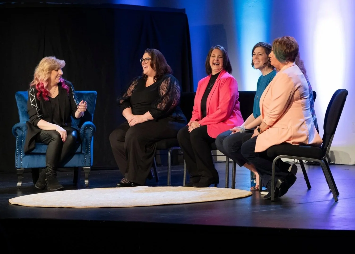Five women sitting on stage, engaged in conversation, with the woman on the far left gesturing as the others listen and laugh. Background is decorated with blue and purple lighting.