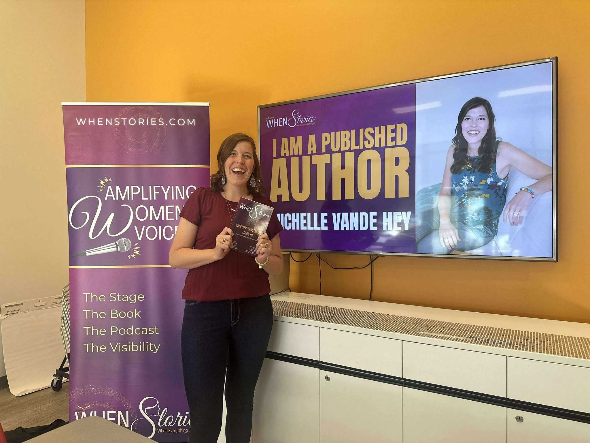 A woman smiling and holding a book standing next to a purple banner and a digital screen displaying the title "I am a Published Author" and her name, Michelle Vande Hey.