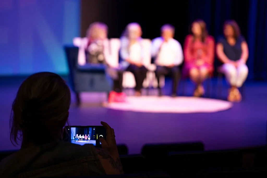 A person taking a photo of a panel of five women sitting on stage in a theater or auditorium.