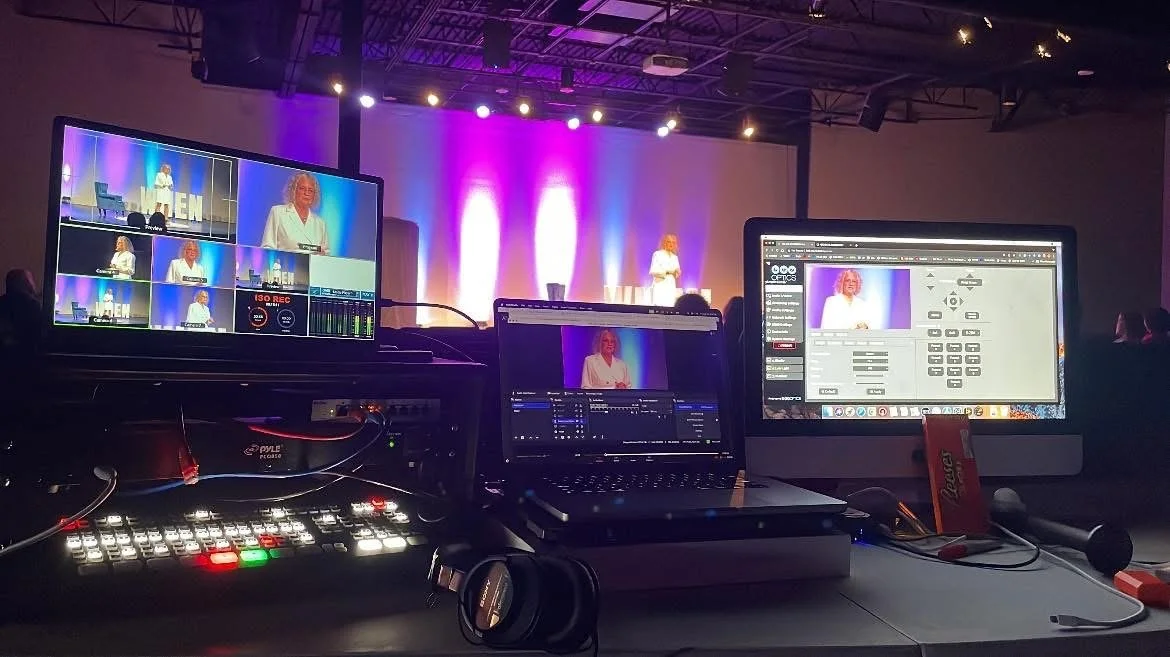 Stage with a woman speaking at a conference, two computer screens and audio equipment in the foreground.