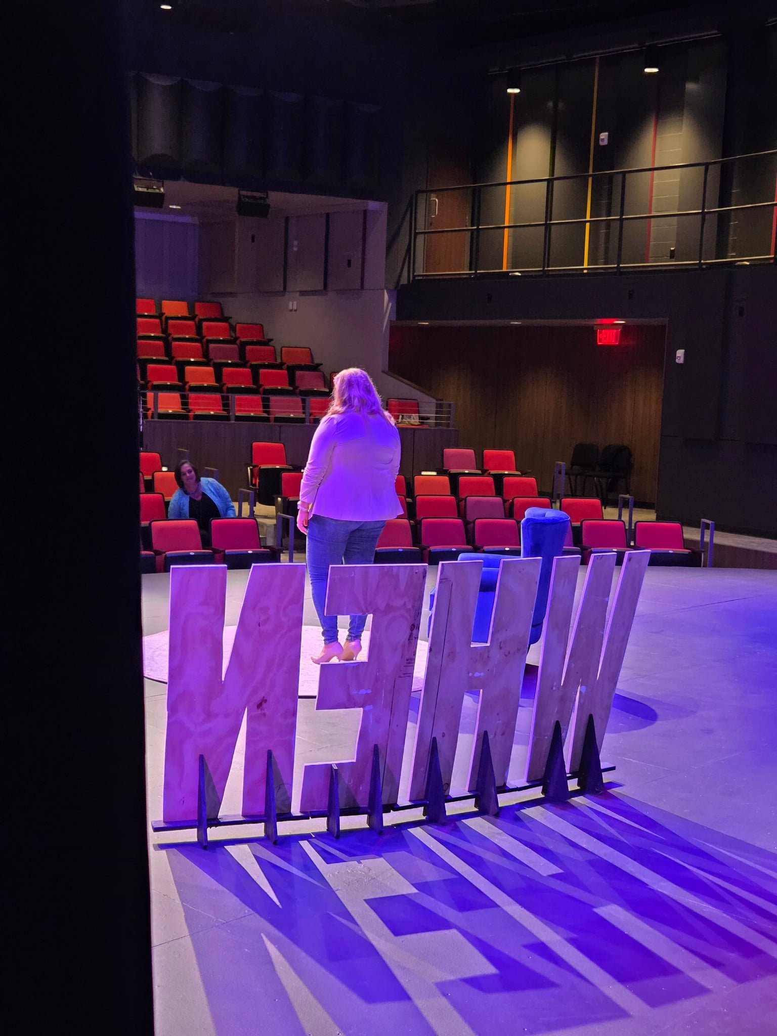 Empty theater or auditorium with red seats and two women standing and talking, illuminated by purple and blue stage lights, with a wooden accent wall and a balcony with black railing.