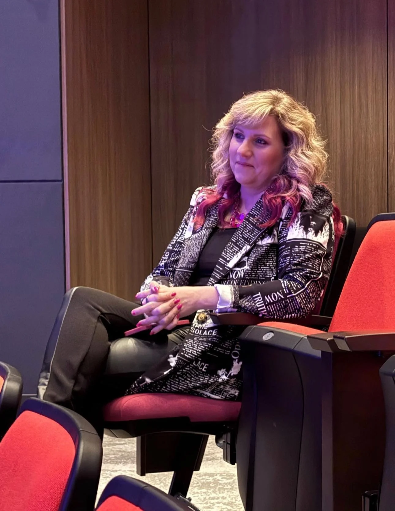 A woman with blonde hair and pink tips, wearing a black and white patterned blazer and black pants, sitting in a row of red chairs in a conference or meeting room with wood-paneled walls.