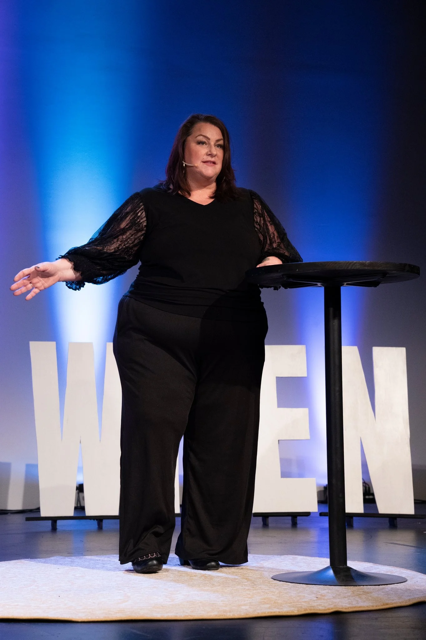 A woman giving a presentation on stage, standing beside a small round table, with a large illuminated sign behind her that reads 'WOMEN'.
