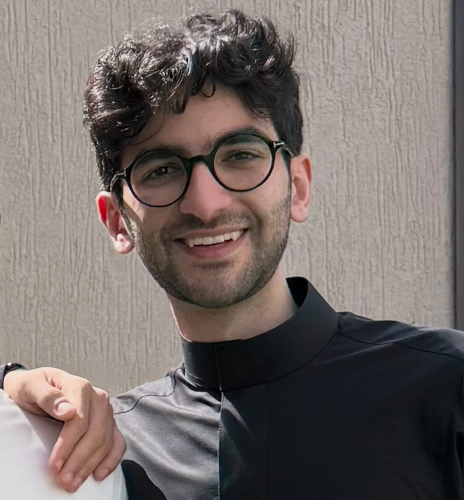Close-up of a young man with dark, curly hair, wearing glasses, smiling, holding hands with another person.