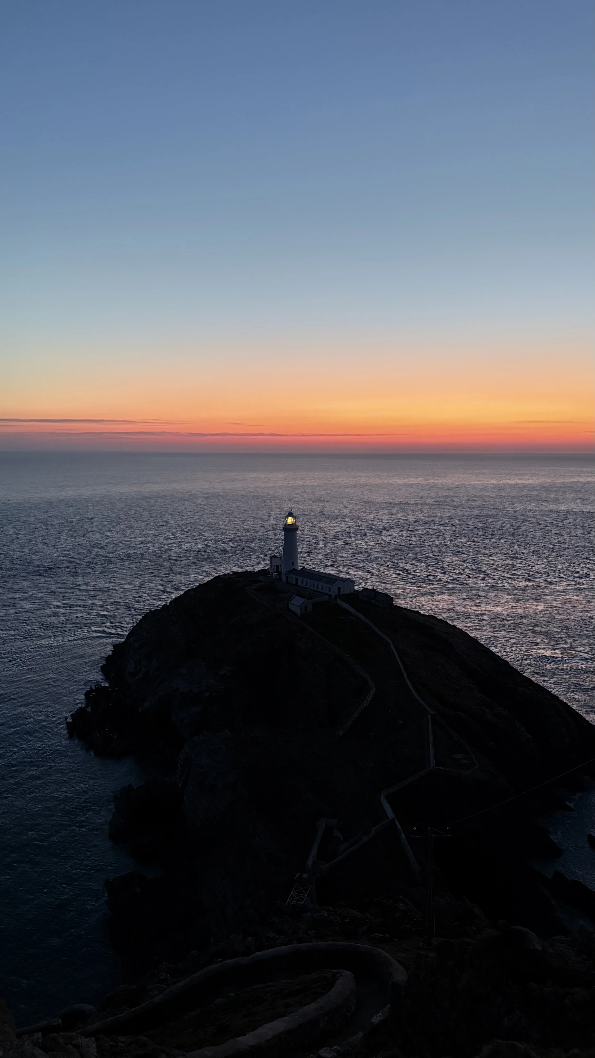 Sunset over the ocean with a lighthouse on a small rocky cliff.