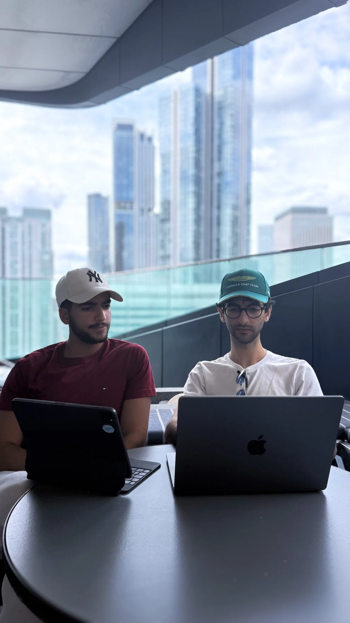 Two young men working on laptops at a table in a modern office with large windows and a city skyline in the background.