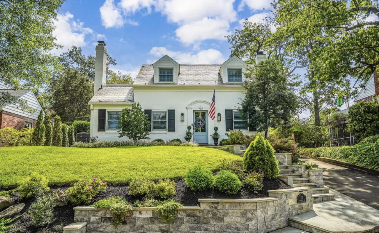 Sylvan white house with a well-maintained front yard and garden, American flag, blue sky with clouds, trees, and surroundings.
