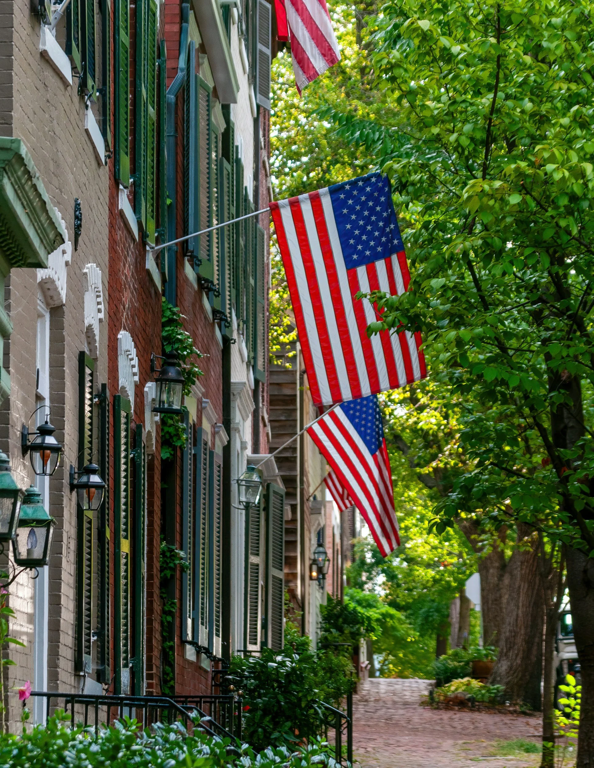 Street scene with brick townhouses decorated with green shutters and American flags, lush green trees lining the sidewalk, and a cobblestone path.
