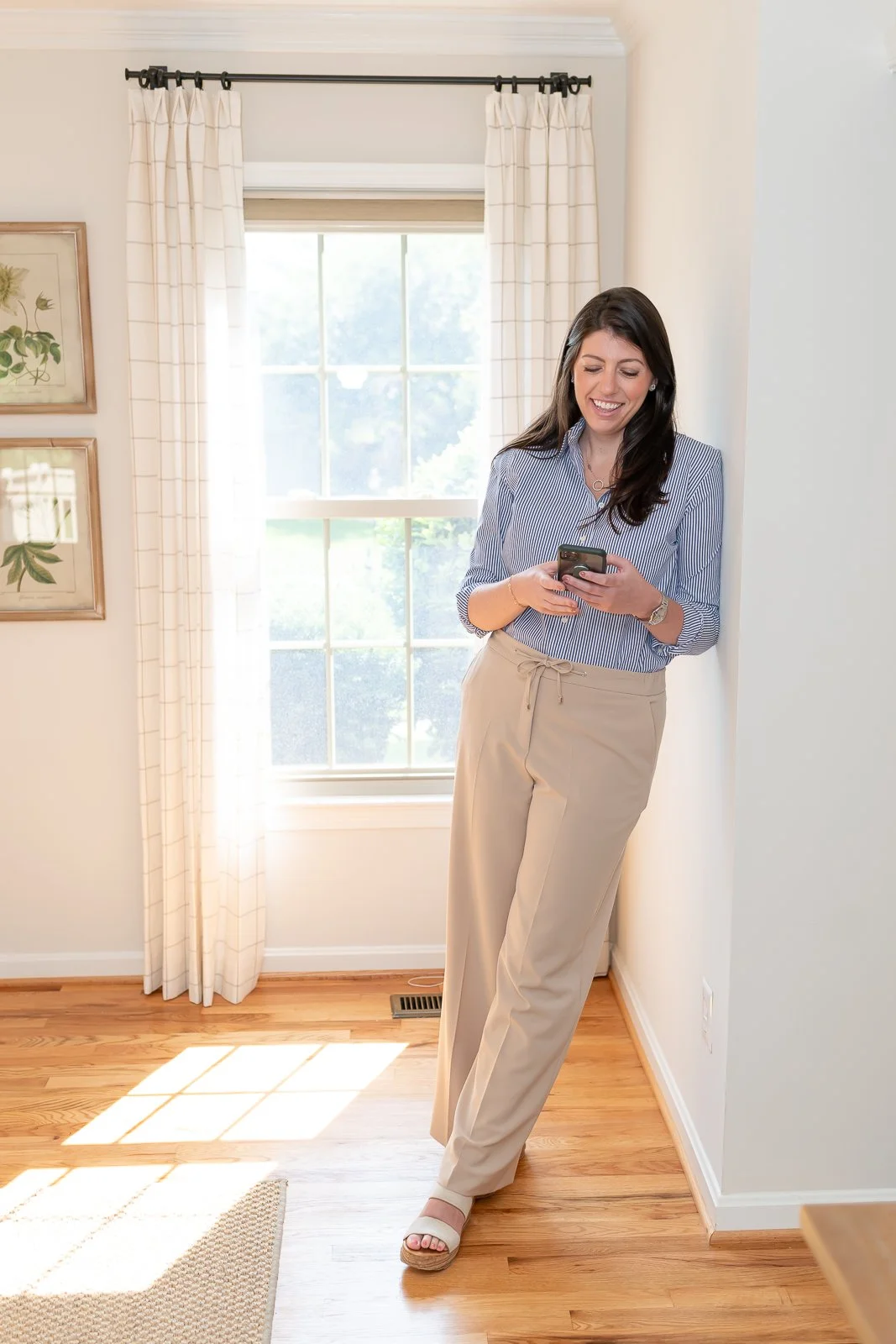 A woman with long dark hair and light skin, wearing a blue and white striped button-up shirt and beige pants, standing in a bright room with a window, looking at her phone, smiling.