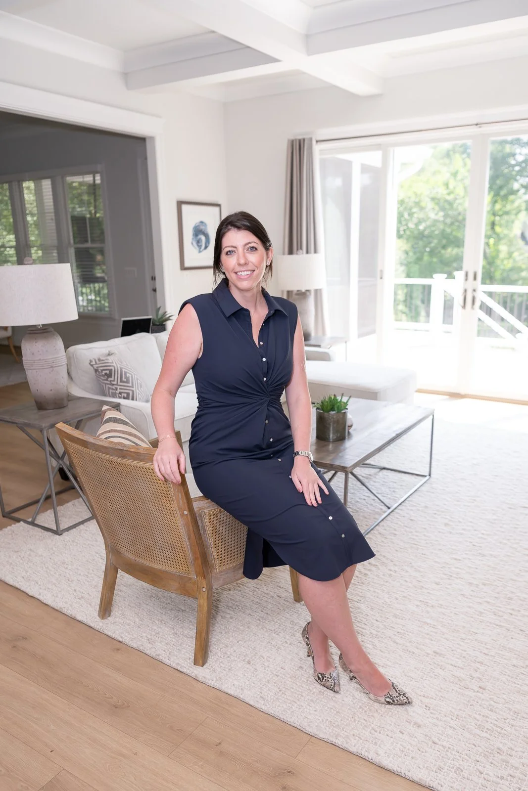 A woman in a navy dress sitting on a wooden chair in a bright, modern living room with a glass door leading outside, a white couch, side tables, and decorative plants.