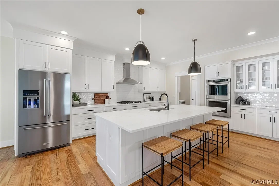 Modern white kitchen with stainless steel refrigerator, white cabinets, and a large island with four woven barstools. Pendant lights hang over the island, and appliances include a range hood and double oven.
