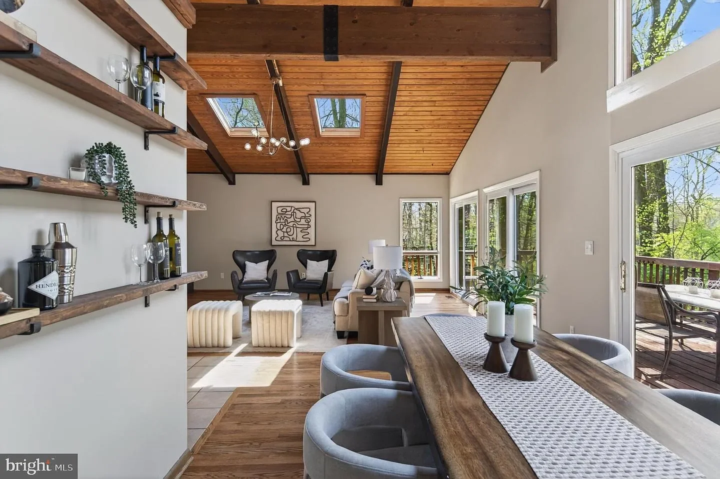 Open living room with vaulted wooden ceiling, large windows, a dining table with candles, a sitting area with armchairs and a sofa, and outdoor deck visible through sliding glass doors.
