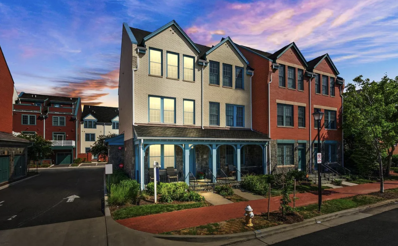 Modern multi-story townhouses with a porch, lit from inside, neighborhood street during sunset, with trees, lampposts, and a sidewalk.
