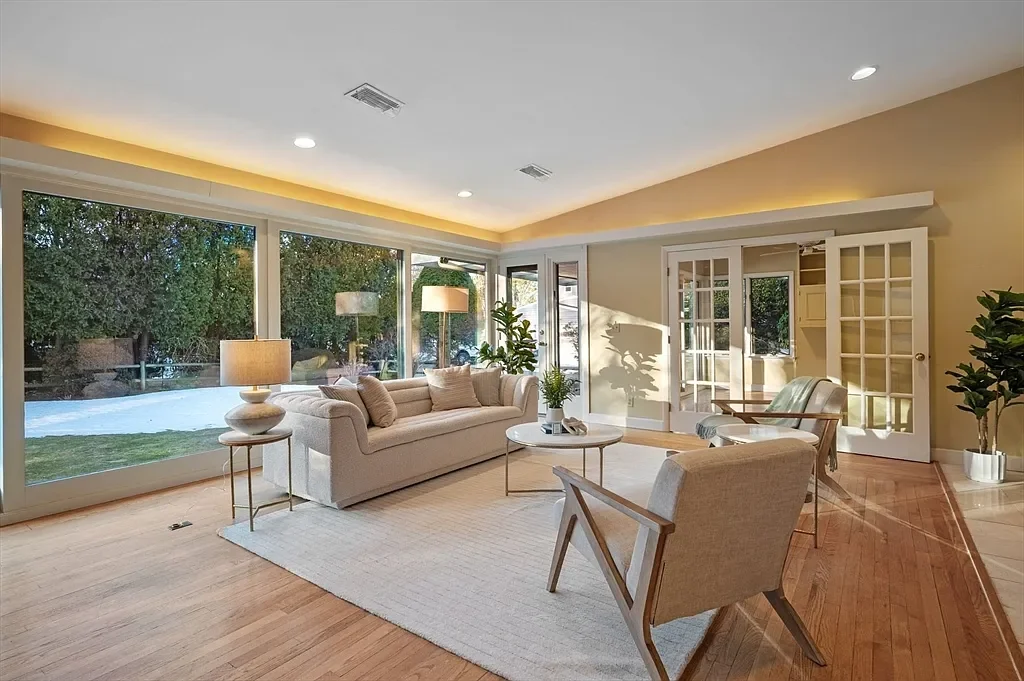 Bright living room with large windows, beige sofa, two armchairs, and a round coffee table. Interior has hardwood floors, plants, and sliding glass doors leading outside.