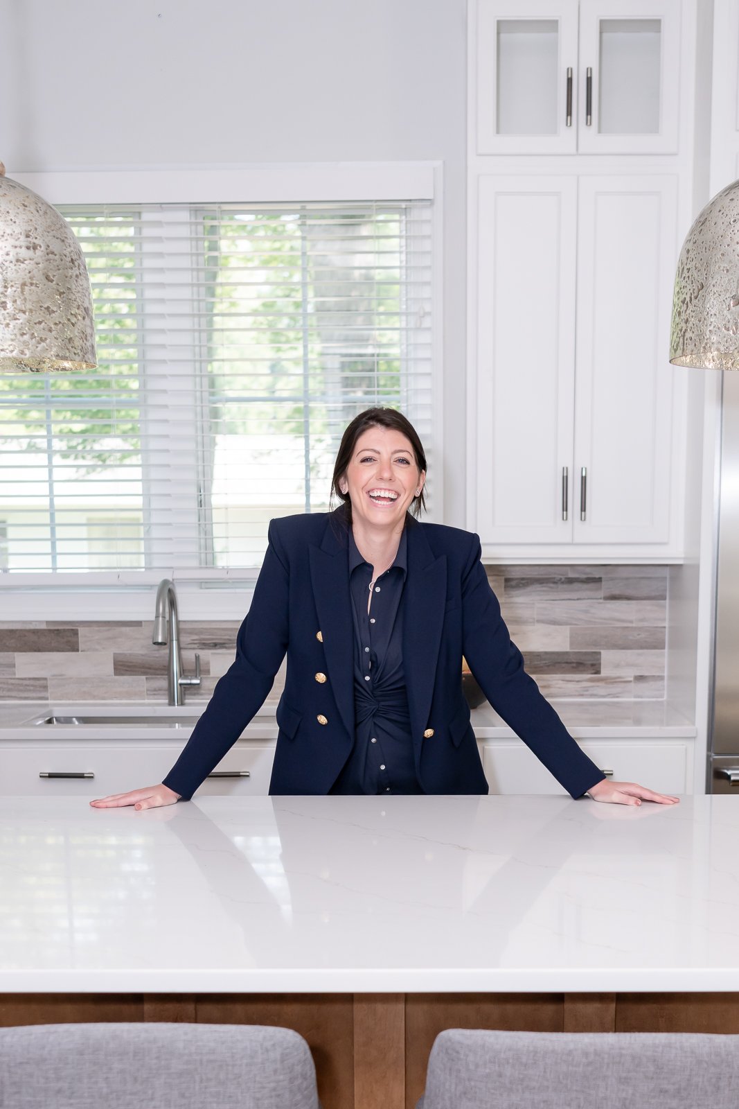 A woman with dark hair, dressed in a dark blue blazer, smiling and leaning on a kitchen counter in a modern kitchen with white cabinets and a large window with blinds.