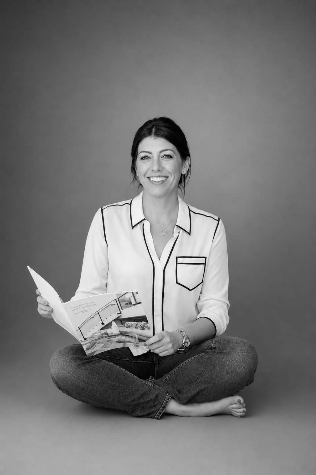 Black and white photo of a woman sitting on the floor with legs crossed, smiling, holding an open magazine, wearing a light-colored blouse with dark piping and dark jeans, against a plain background.