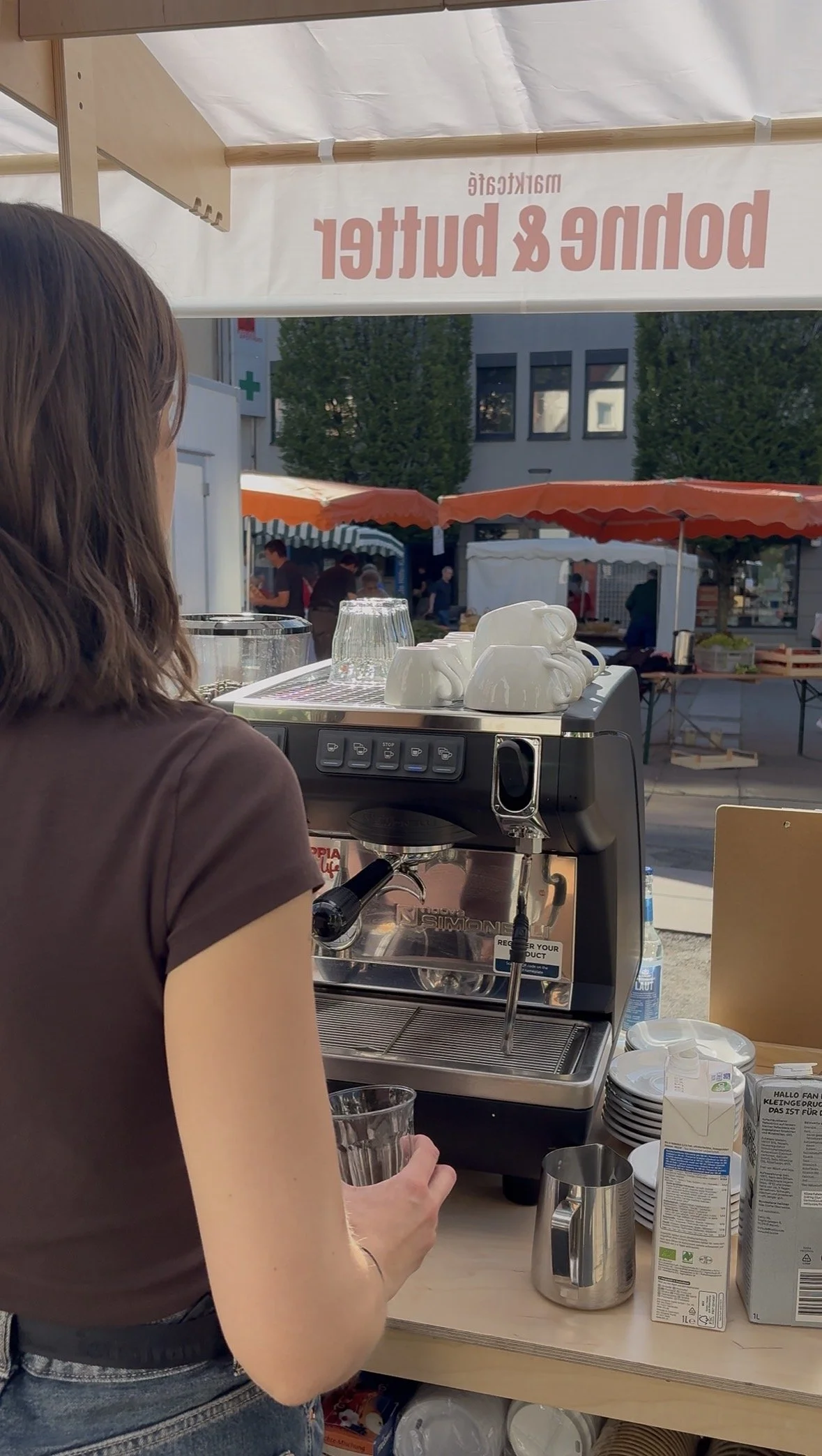 Eine Person bereitet Kaffee mit einer Espressomaschine zu an einem Stand im Freien, im Hintergrund sind Marktzelte und Bäume sichtbar.