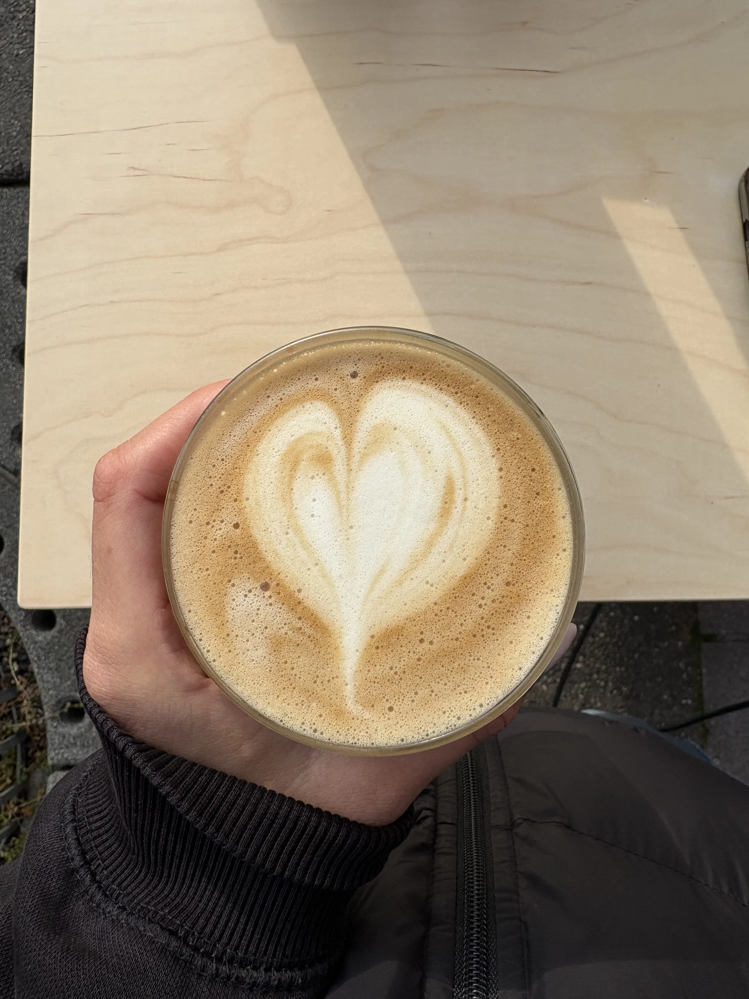 Eine Hand hält einen Kaffee mit Herz-Muster auf der Oberfläche. Der Hintergrund zeigt eine helle Holzfläche.