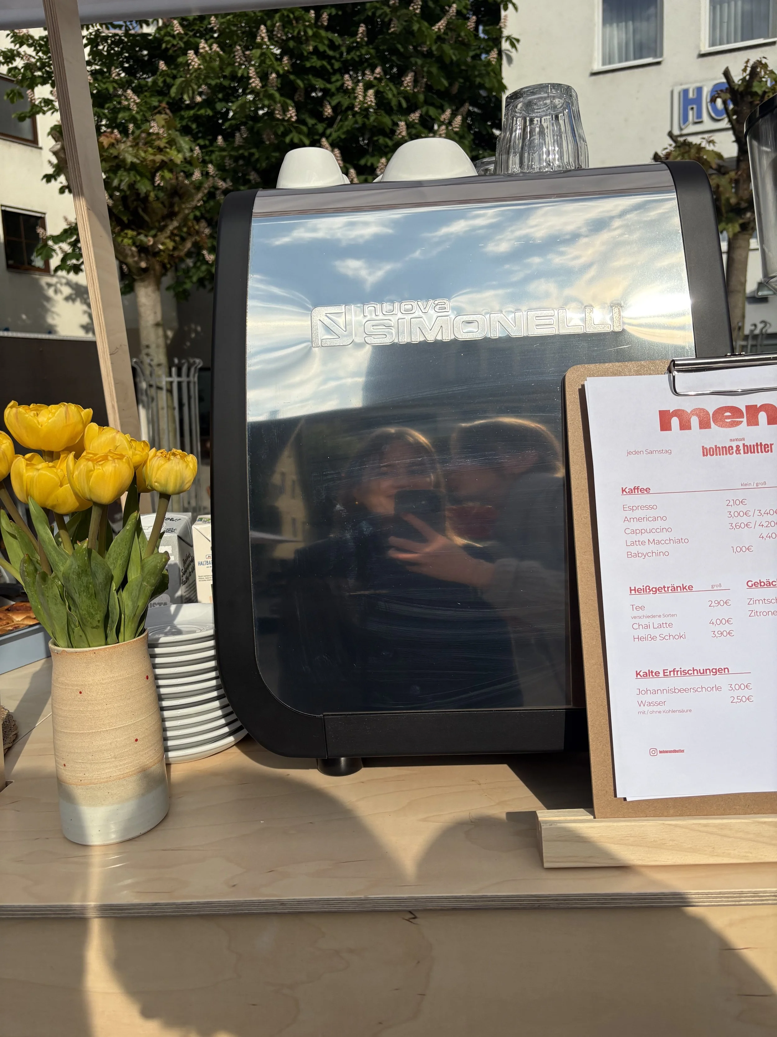 Ein Automat für Getränke und Kaffee, mit einer Tafelmenü, einer Vase mit gelben Tulpen und einem Blick auf den Himmel im Hintergrund.