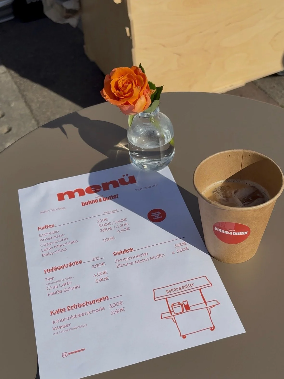 Ein Tisch mit einer rosafarbenen Blume in einer kleinen Vase, einer Tüte Kaffee mit Eiswürfeln und einem Menüblatt mit Speisen und Getränken, auf dem Tisch. Das Menü ist von einem Café namens "bone & butter".