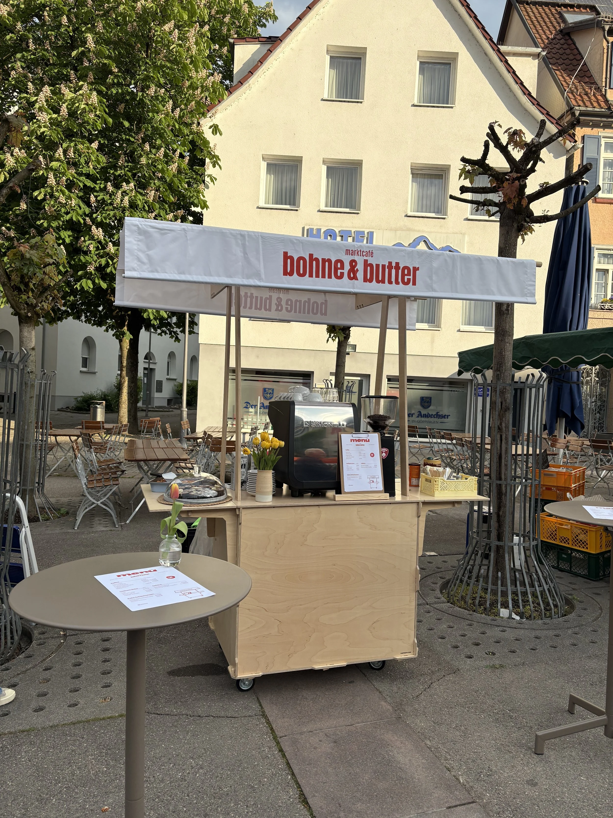Straßenverkaufsstand für Kaffee und Gebäck mit Tischen und Stühlen im Außenbereich, Bäume und Gebäude im Hintergrund.