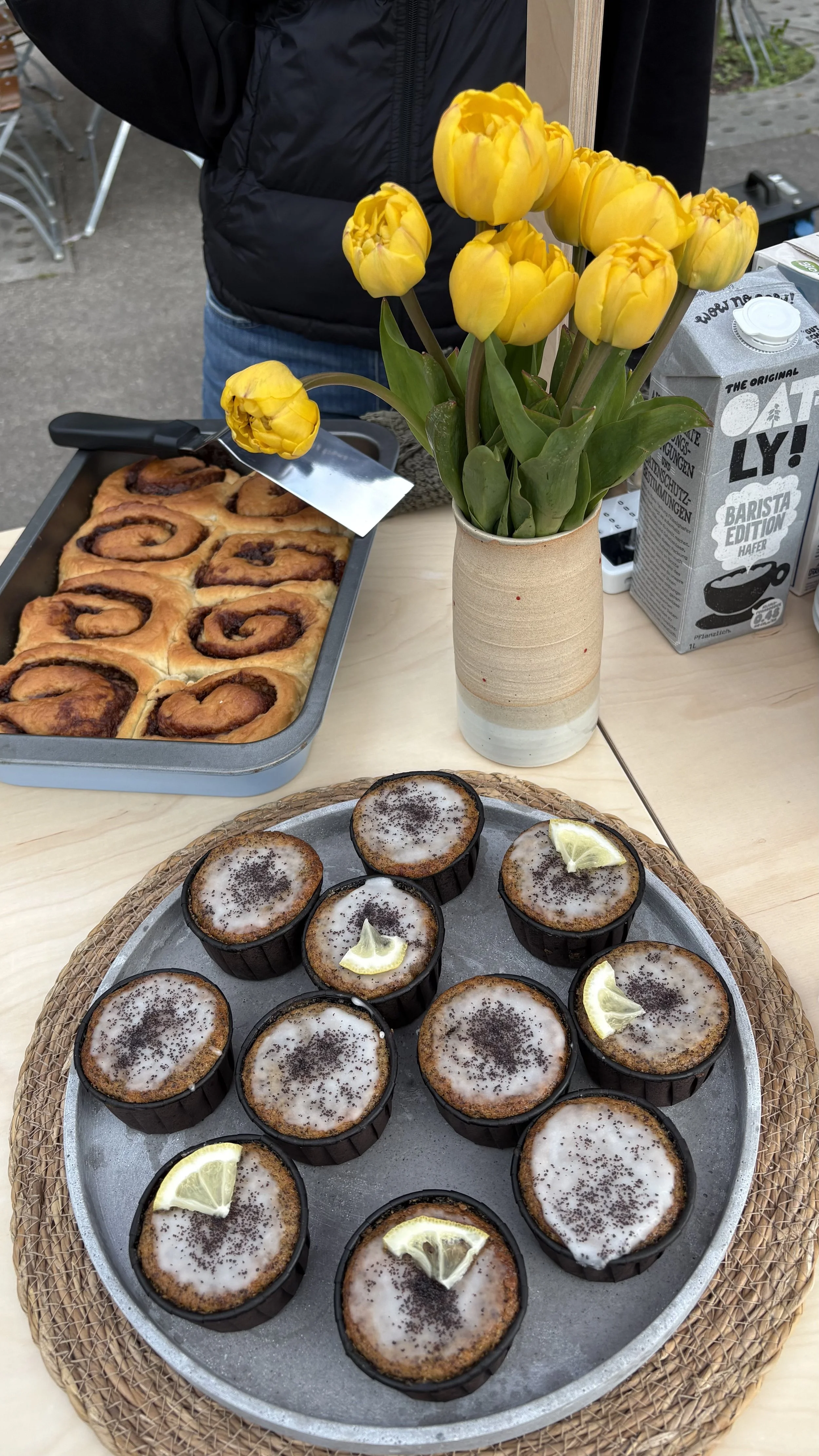 Tassen mit Kuchen, eine Vase mit gelben Tulpen, eine Backform mit Zimtschnecken und eine Packung Hafermilch auf einem Tisch.