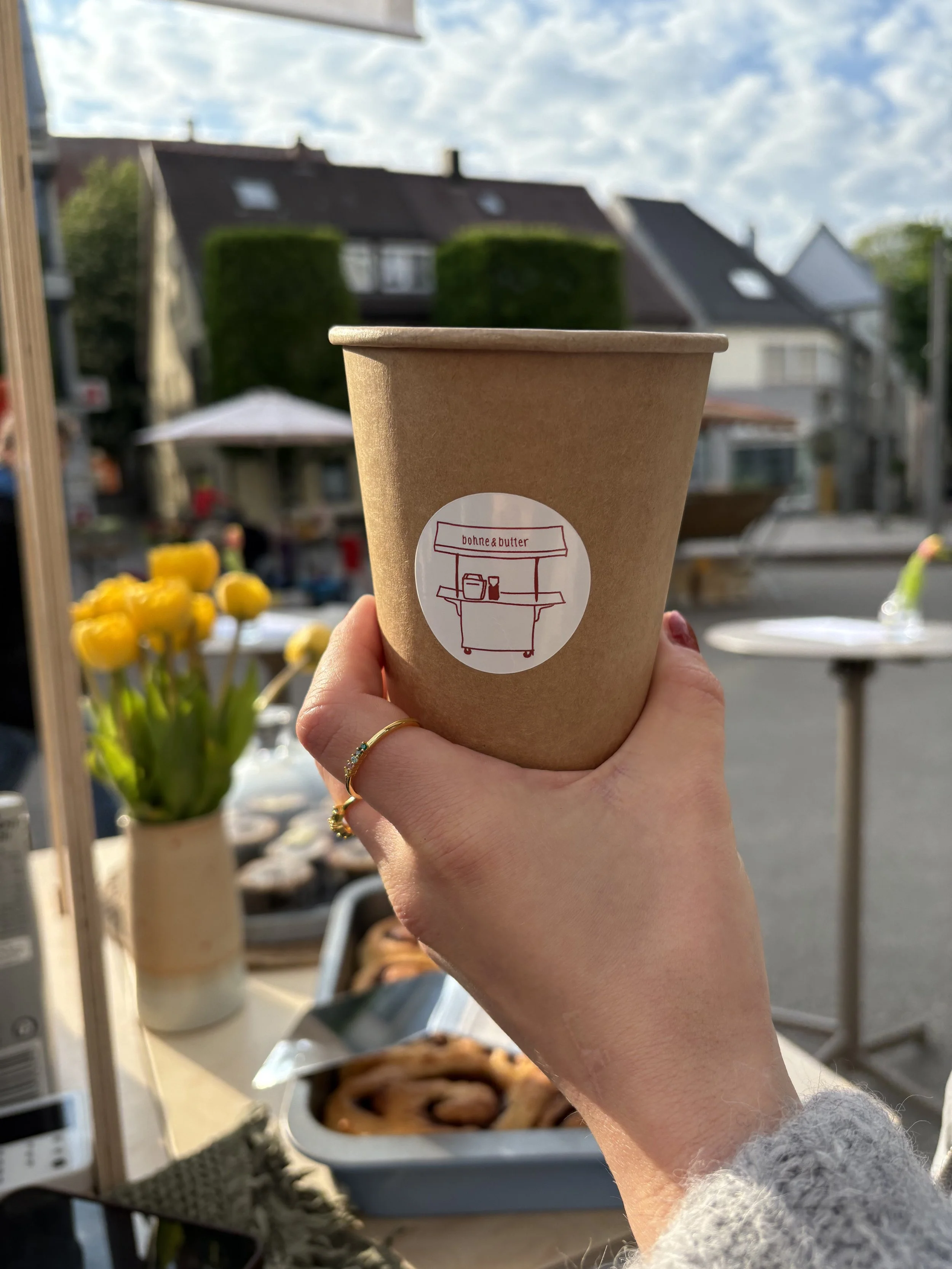 Eine Hand hält eine braune Pappbecher mit einem Aufkleber, auf dem eine Straßenverkäufer-Illustration und der Schriftzug "bonne & butter" sind, im Freien bei sonnigem Wetter. Im Hintergrund sind Häuser, ein Blumenmarkt mit gelben Tulpen, und ein blau