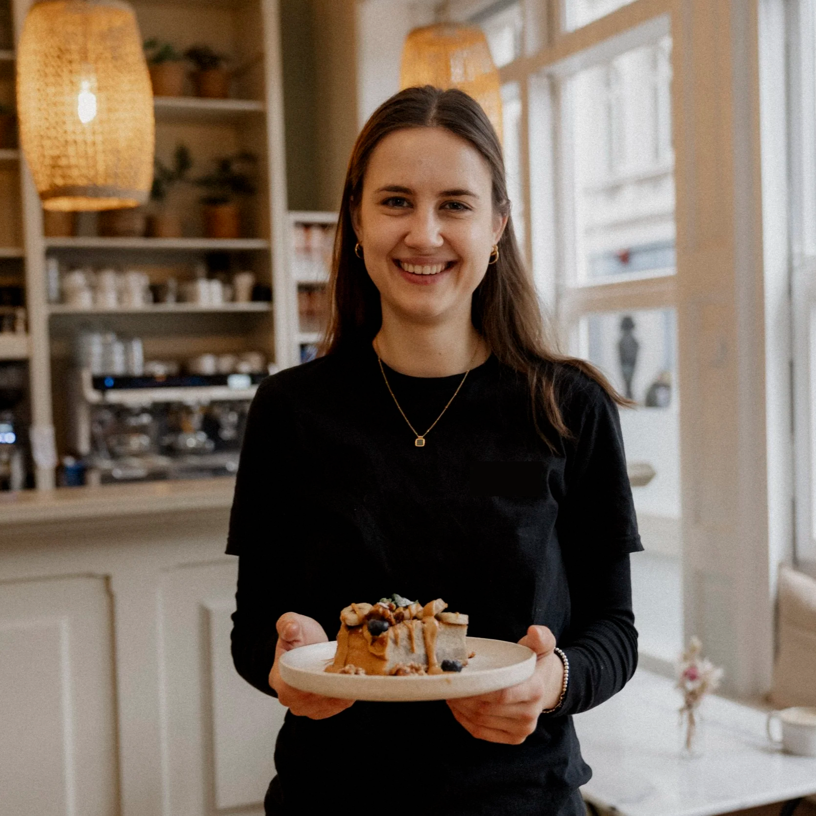 Eine junge Frau hält einen Teller mit Kuchen im Wohnzimmer, im Hintergrund sind Lampen, Bücherregale und Fenster sichtbar.