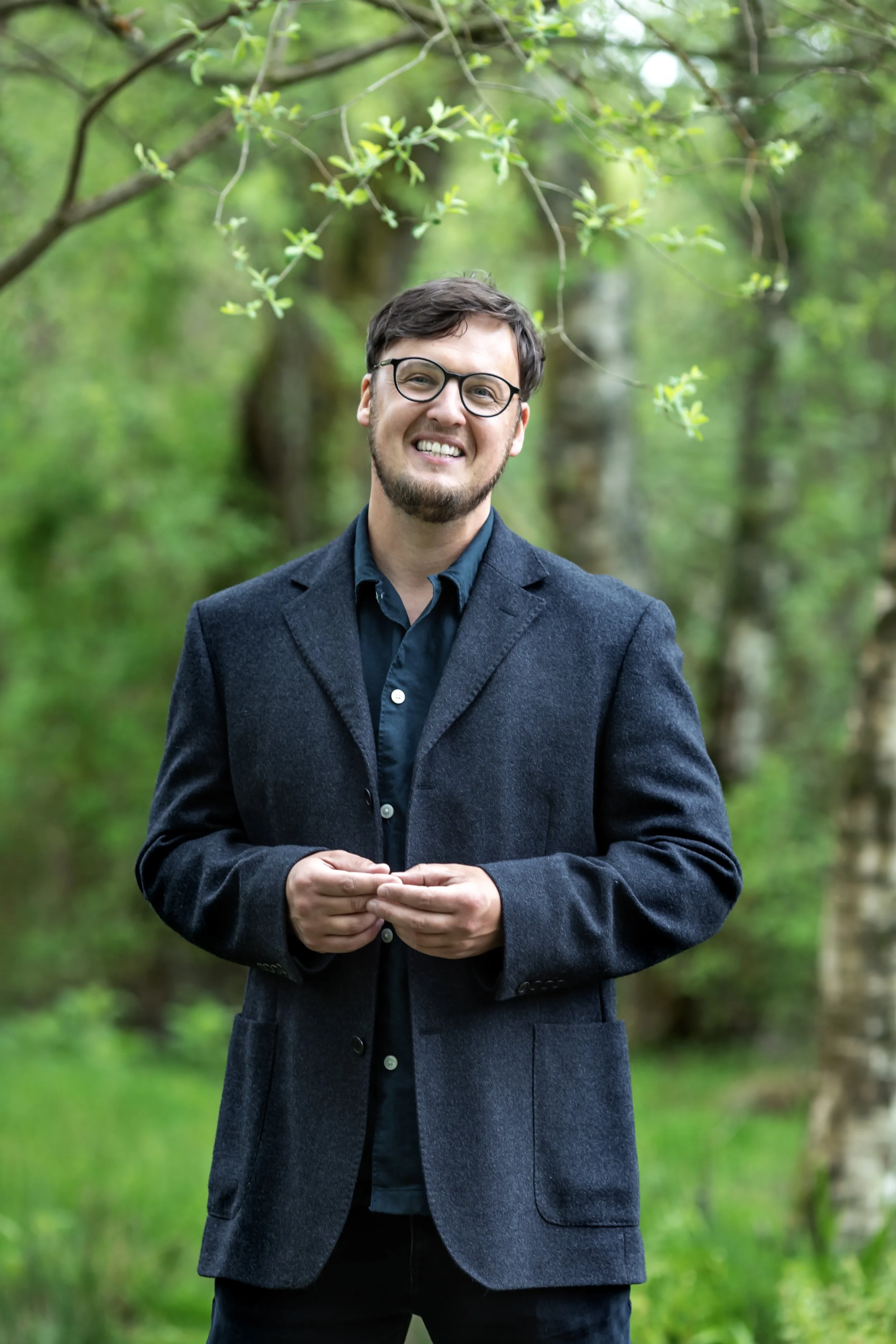 A man with dark hair, glasses, and a beard wearing a dark blazer and a blue shirt, smiling and standing outdoors among green trees.
