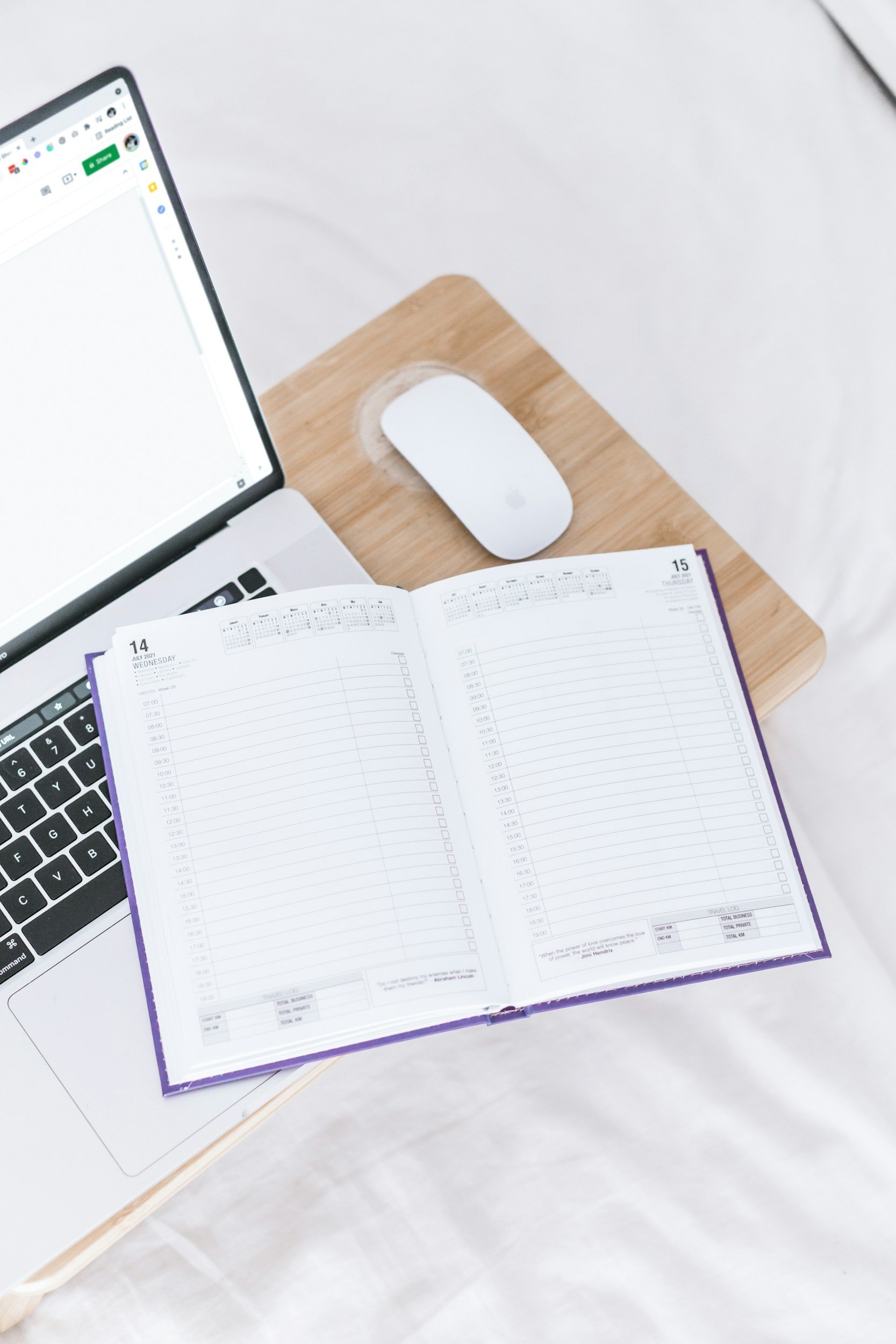 Open planner with weekly schedule, closed laptop, white computer mouse, and a laptop on a wooden table.