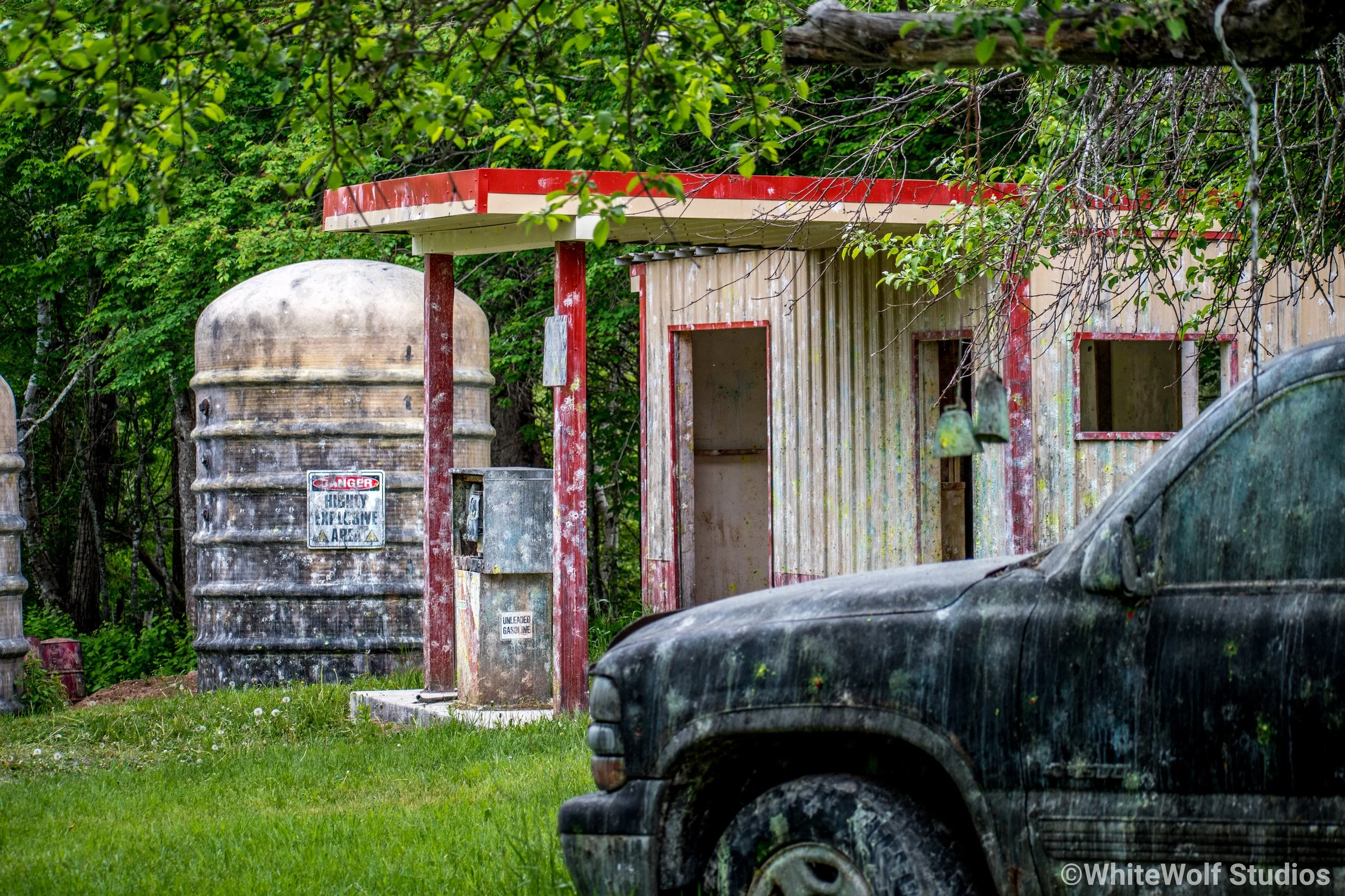 An overgrown, abandoned gas station with rusted tanks, weathered wooden structures, and old cars covered in dirt, surrounded by green trees.