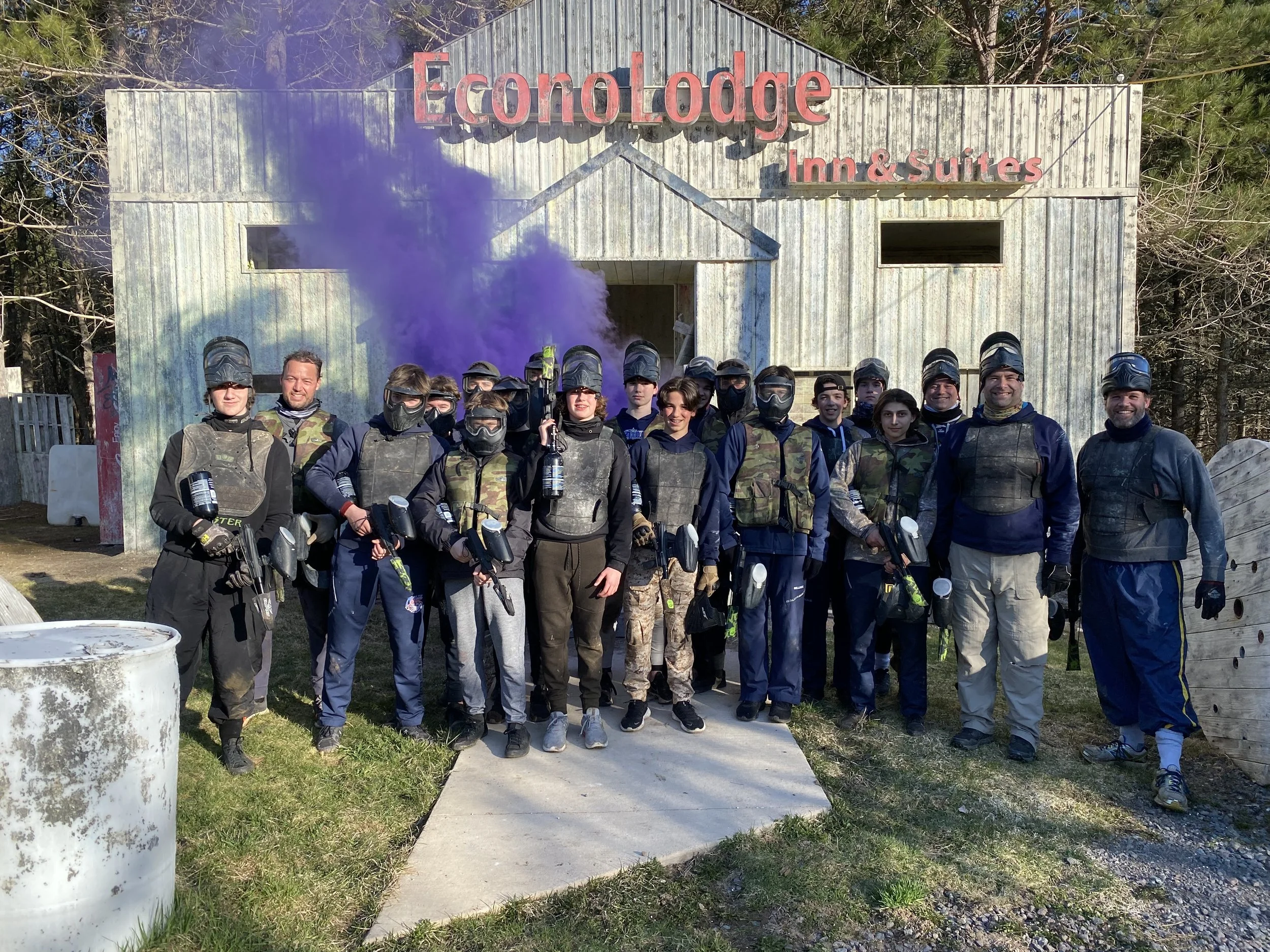 A group of people dressed in paintball gear, holding paintball guns, standing outside in front of a sign that reads 'Ecologde Inn & Suites', with trees and a weathered building in the background.