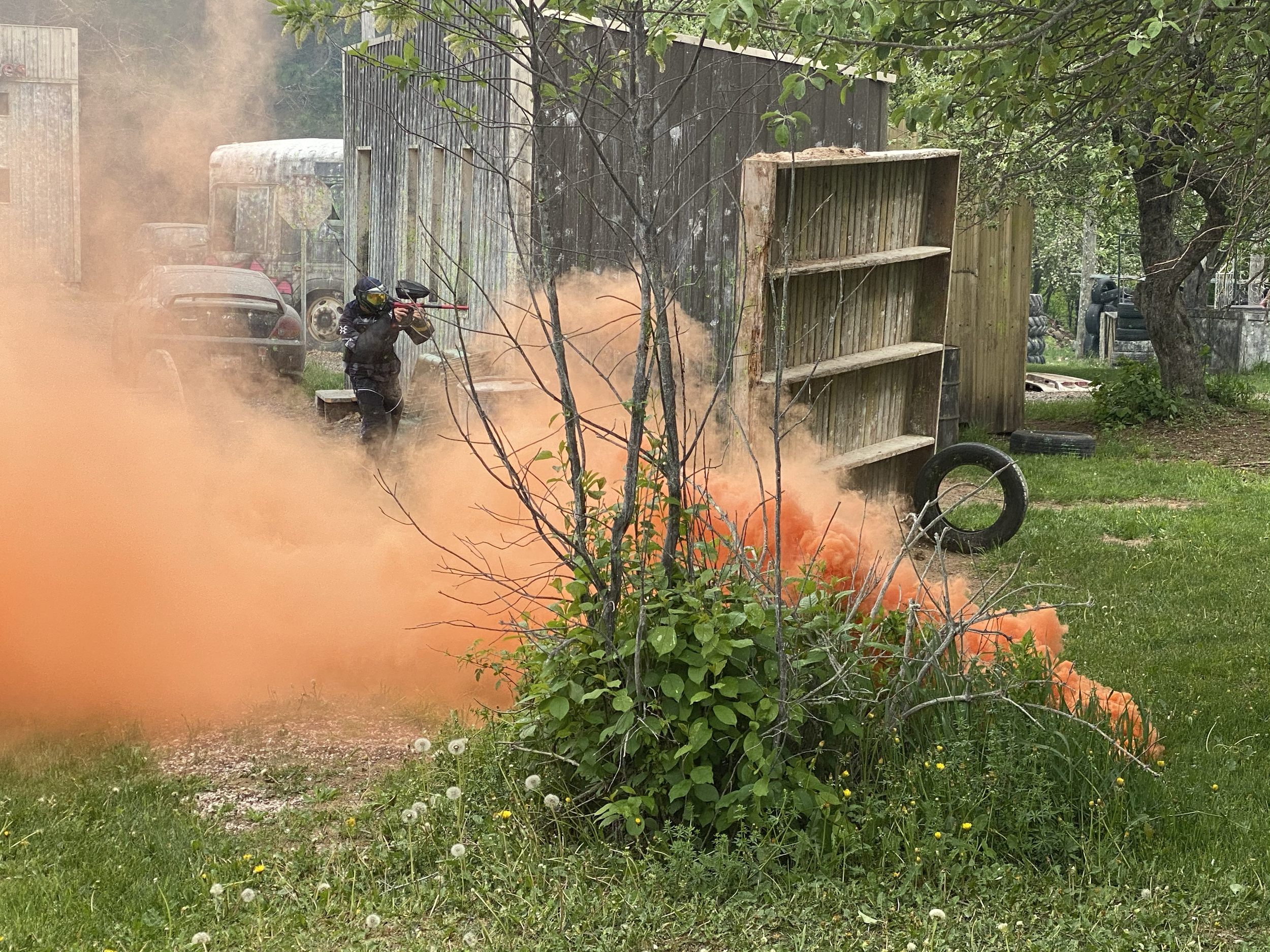 A person wearing a paintball mask and holding a paintball gun behind orange smoke on a paintball field. There are wooden structures, tires, and old vehicles in the background, surrounded by trees and grass.