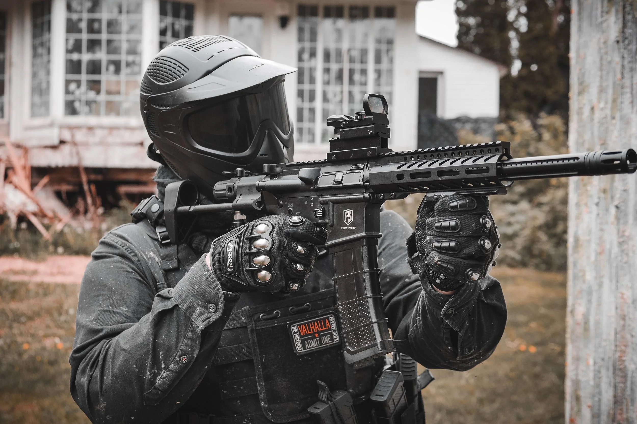 A person wearing gloves and tactical gear holding a rifle with a helmet resting on their shoulder, outdoors in front of a house with large windows.