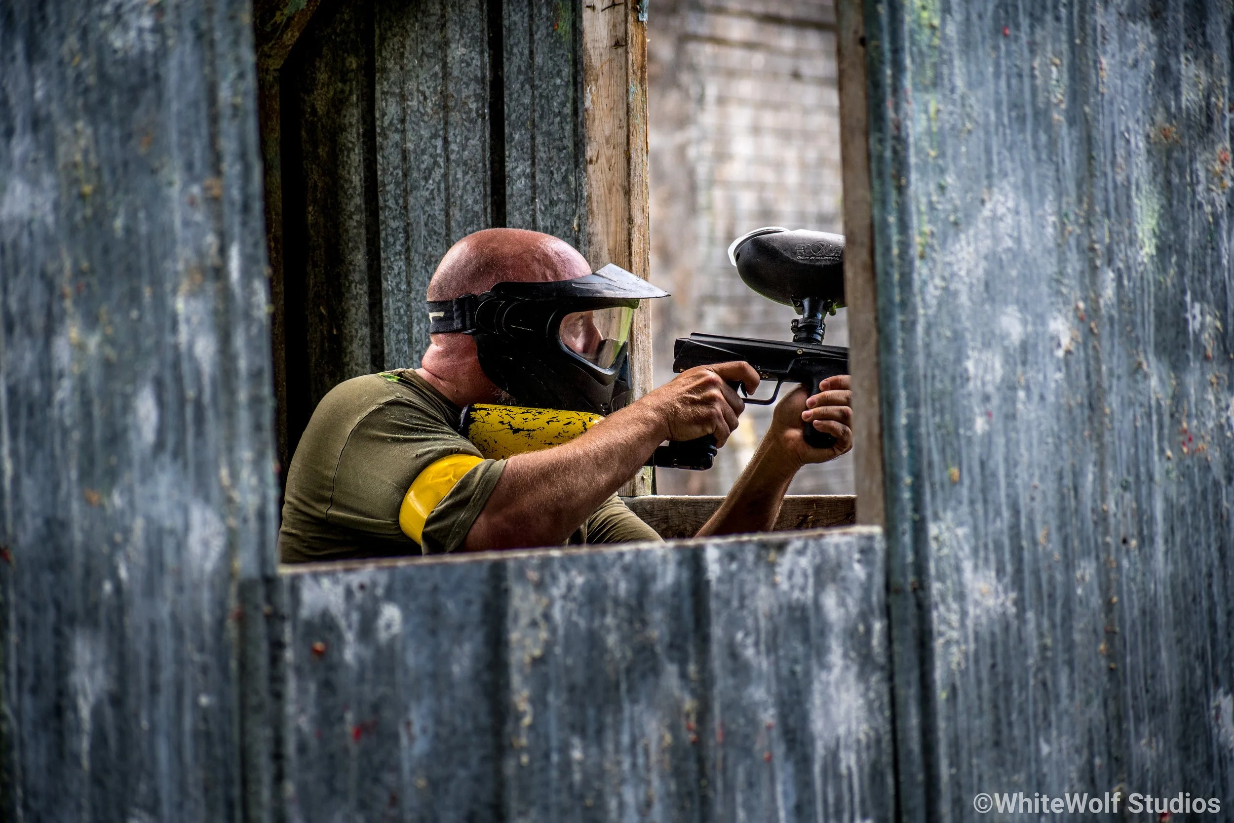 A person wearing a helmet and protective gear aiming a paintball gun through a narrow opening in a weathered wooden and corrugated metal structure during a paintball game.