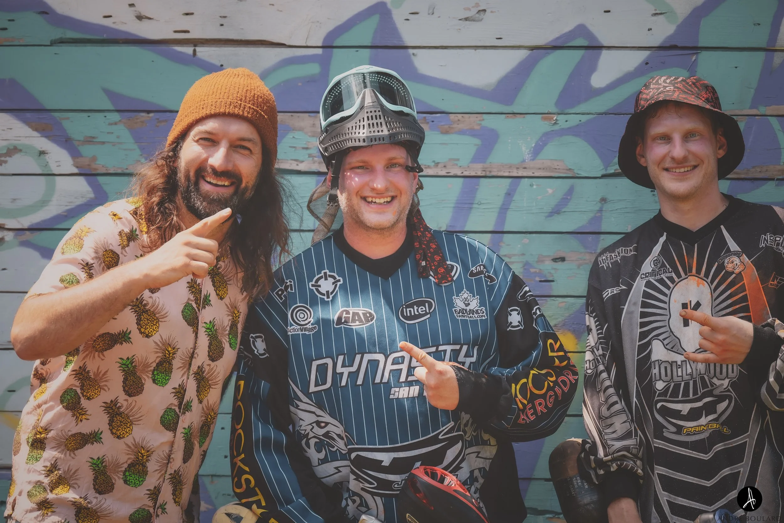 Three men smiling and pointing at the camera, standing against a colorful, graffiti-covered wooden wall. The man in the middle is wearing a full face paint paintball mask on top of his head and a dark blue paintball jersey with various sponsor logos.