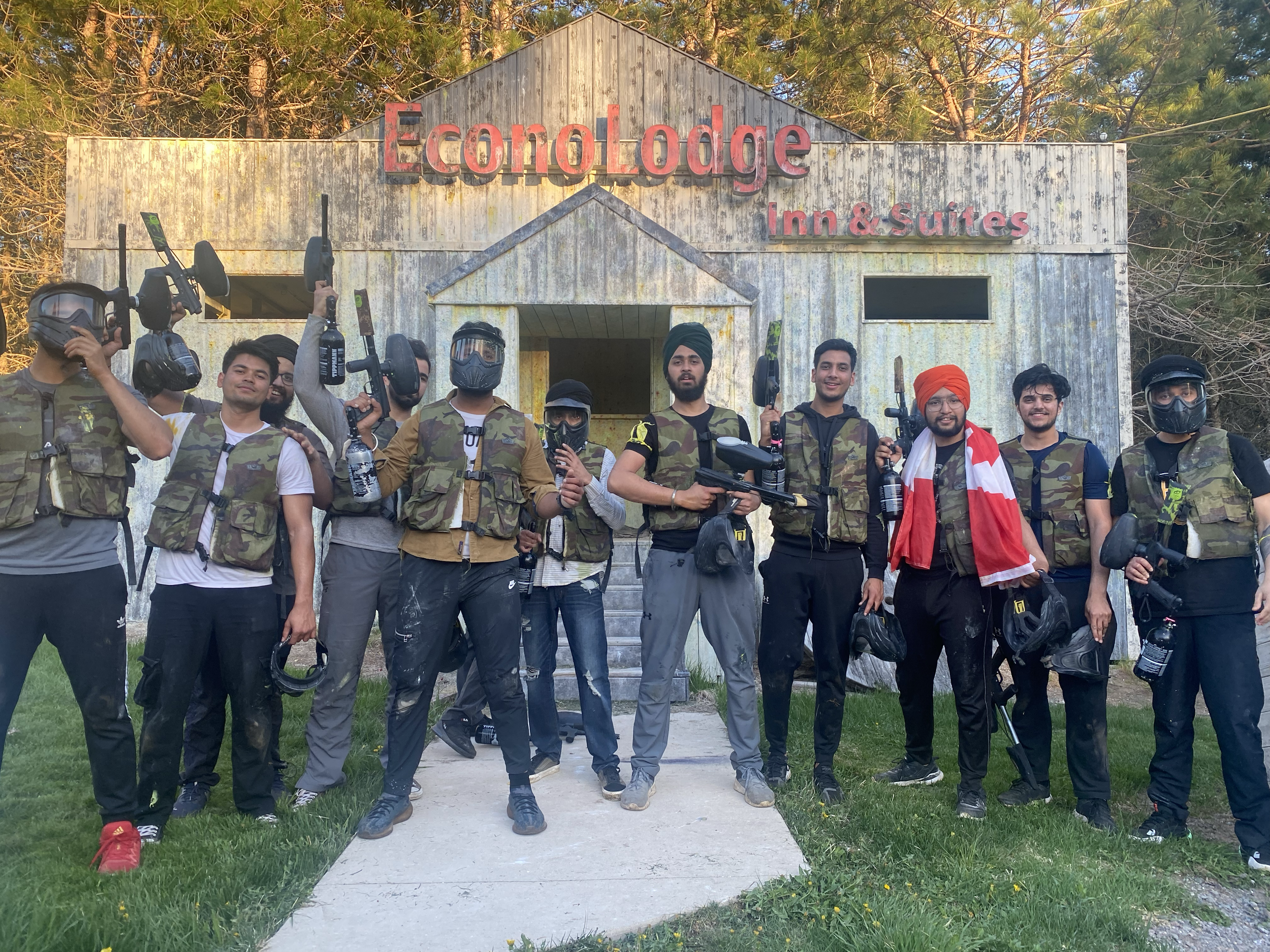 A group of ten young men, some wearing camouflage vests and helmets, posing with paintball guns in front of a rustic wooden building with a sign that reads 'EconoLodge Inn & Suites' surrounded by trees.