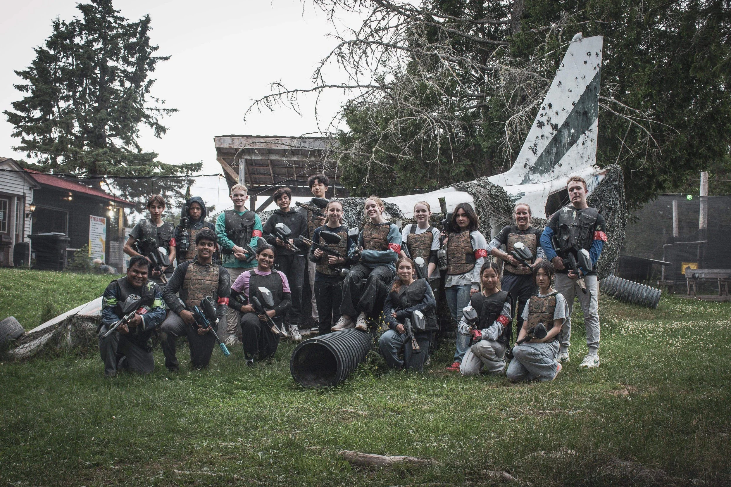 A group of children and teenagers posing outdoors in front of a damaged airplane fuselage with foliage in the background, some holding paintball guns and wearing protective gear.