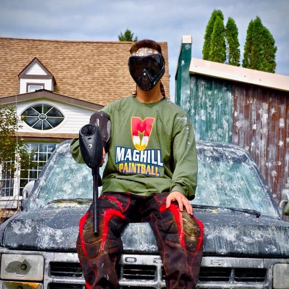 A person wearing a paintball mask, holding a paintball marker, and sitting on the hood of an old, dirty car. The person is wearing a green sweatshirt with the text 'Maghill Paintball' and camouflage paintball pants. The scene is outdoors, with a hous