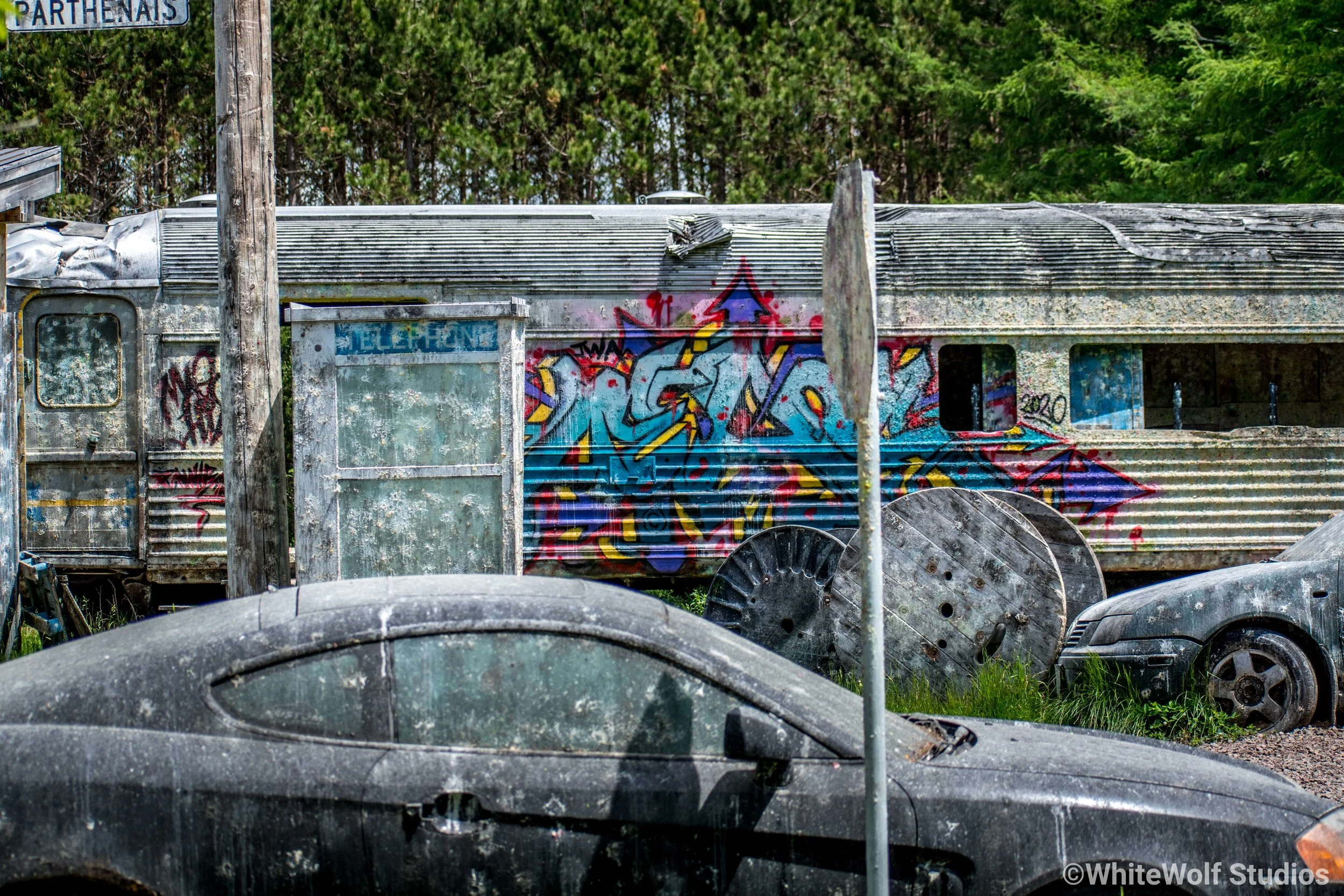 Old, weathered mobile home with graffiti, surrounded by trees and old car tires in the foreground.
