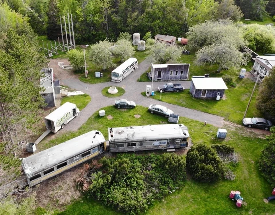 An aerial view of a rural area with old train cars, several cars, small buildings, trees, and grassy areas.