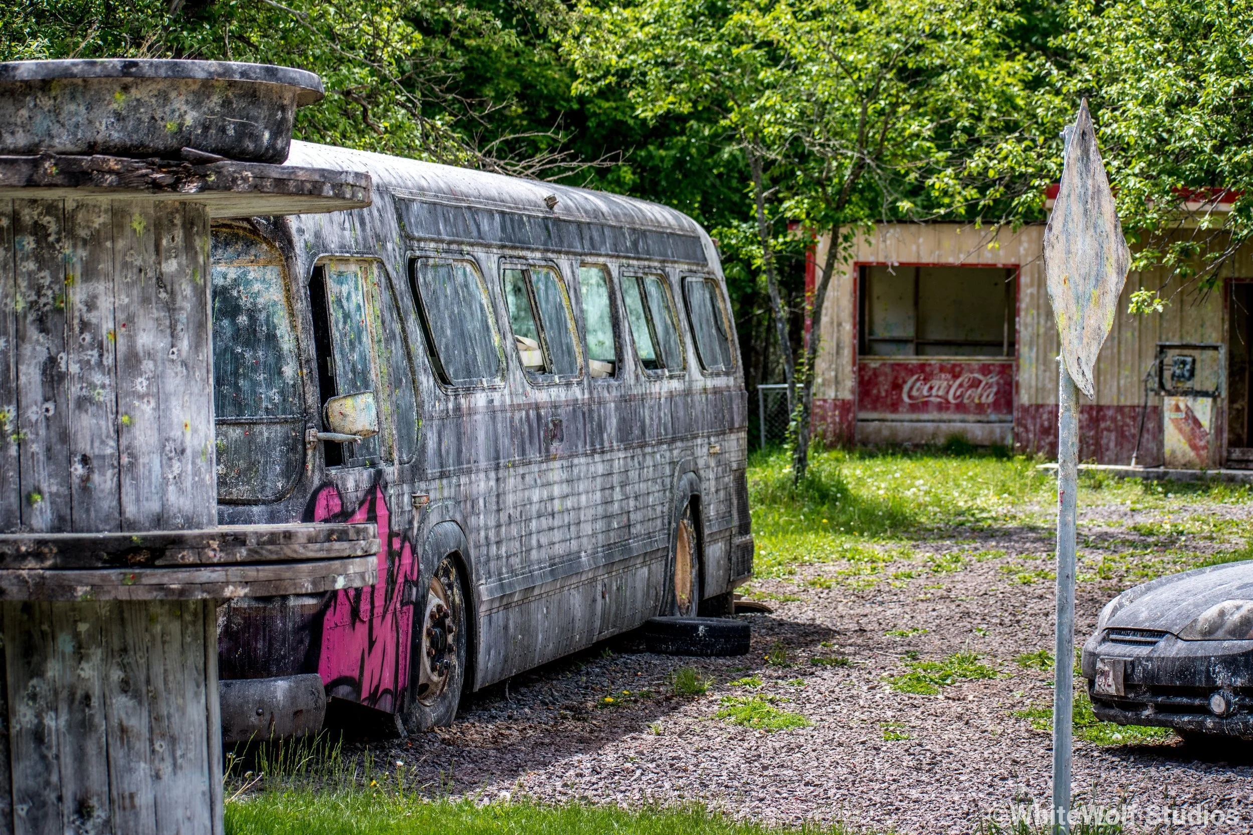 An abandoned, weathered bus covered in dirt and graffiti parked near a small building with a Coca-Cola sign, surrounded by trees and grass.